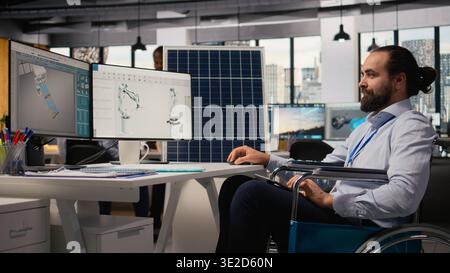 Man with paraplegia in inclusive RD office designs green technology solar panels. Employee with disability in wheelchair runs simulation to enhance solar cells, clean energy production, camera A Stock Photo