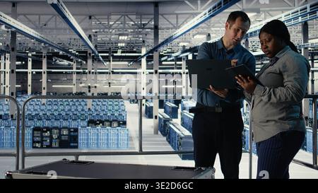 Team of engineers monitor server hub activity using laptop and tablet. Teamworking data center colleagues review rigs tests results on devices to ensure systems are working correctly, camera A Stock Photo