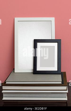 Large white and small black photo frames with mat board leaning on a stack of books against dusty pink wall. Natural side light. Art print mockup for Stock Photo