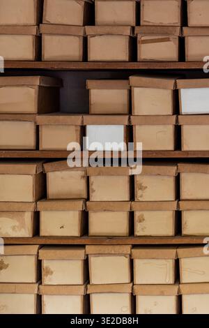 Aged cardboard boxes stacked on dark wooden shelves in a vintage archive warehouse. Warm light, geometric repeating pattern, one empty slot and white Stock Photo