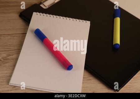 Two markers are lying beside a notepad on a wooden table while a black folder sits in the background. The scene shows tools for writing or drawing. Stock Photo