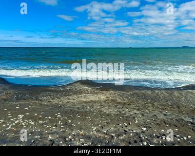 Water meets sand as waves break on the shore, revealing scattered shells. The sky above is blue with clouds, indicating a clear day at the beach in th Stock Photo