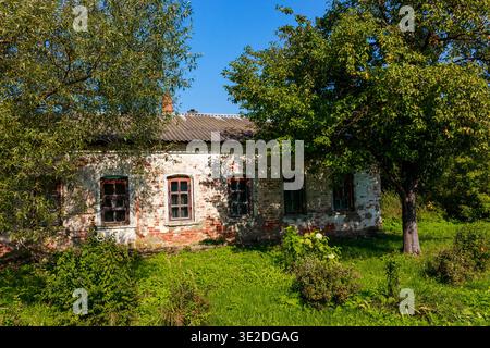 Rustic brick dwelling, its facade peeling, stands amidst vibrant green grass and leafy trees on a sunny day. A quiet, forgotten homestead Stock Photo