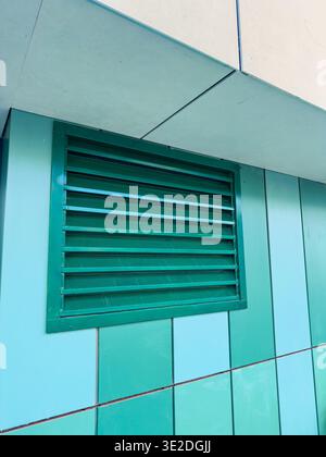 A green vent is mounted on the wall of a modern building. The wall features panels in different shades of blue. Sunlight illuminates the scene. The se Stock Photo
