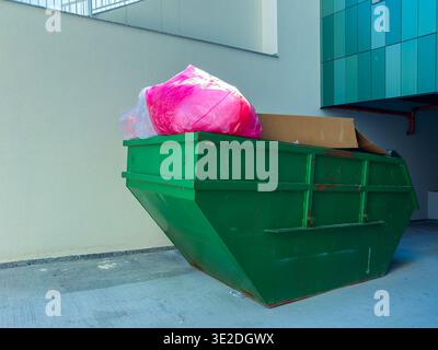 A green dumpster sits next to a building with a pink bag and cardboard on top. It is a clear day, and the wall behind is light-colored and smooth. Stock Photo
