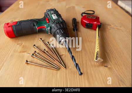 Power drill, wood screws, a screwdriver, and a tape measure are arranged on a rustic wooden work surface, ready for a DIY project. Stock Photo