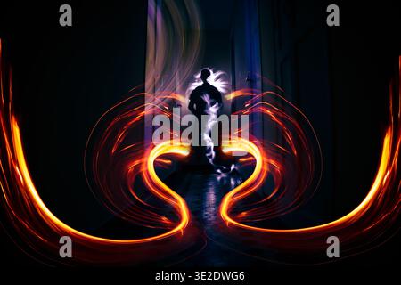 Adult silhouette standing in dark hallway surrounded by swirling red, orange and yellow light trails in long exposure, selective focus. Lightpainting Stock Photo