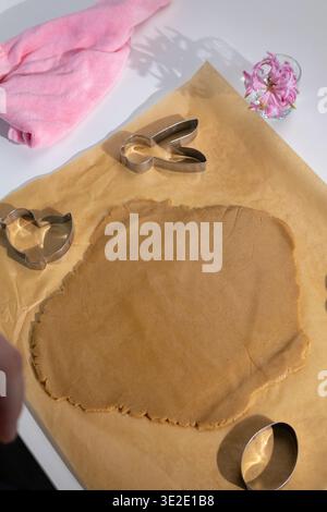 Rolled gingerbread dough sits on parchment paper, ready for cookie cutters. An Easter bunny cutter, a chick cutter, and an oval cutter are visible, alongside a pink towel and pink flowers. Stock Photo
