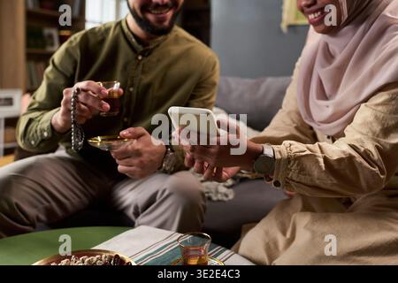 Young Muslim Middle Eastern man holding prayer beads and tea, sitting beside young adult Muslim woman in hijab smiling and showing smartphone screen while sharing moment indoors Stock Photo