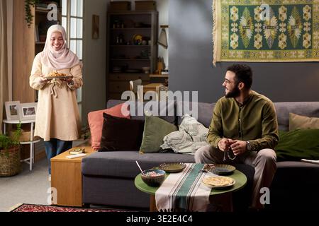 Young Muslim woman wearing hijab carrying plate of food and smiling, young adult Muslim Middle Eastern man sitting on sofa holding prayer beads and looking at her Stock Photo