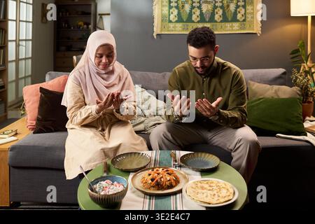 Young adult Muslim woman wearing hijab and young Muslim man sitting on sofa, praying with hands raised before meal on table in cozy living room Stock Photo