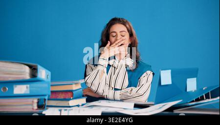 Laptop, files and businesswoman with yawn in studio for filing workload, low energy or fatigue. Tech, folders and tired admin assistant on blue Stock Photo