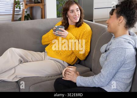 Diverse female friends in mustard knit sweater and gray hoodie sipping mugs on gray sectional couch Stock Photo