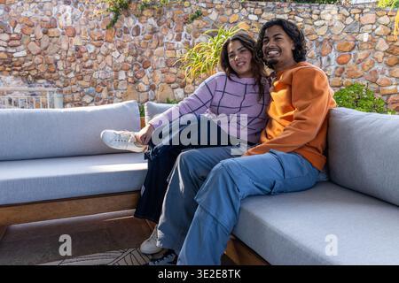 Indian couple sitting closely on backyard patio gray sectional sofa, smiling and holding hands Stock Photo