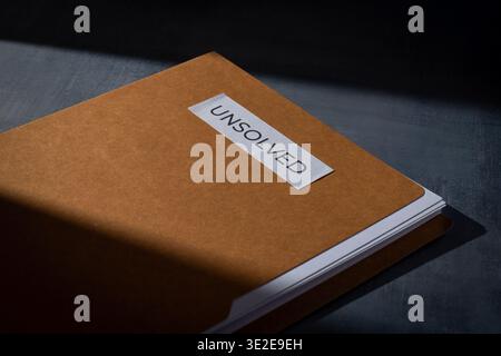 Brown manila folder with UNSOLVED label on a dark surface. Mystery, cold cases, criminal investigation, and unresolved problems concept. Stock Photo