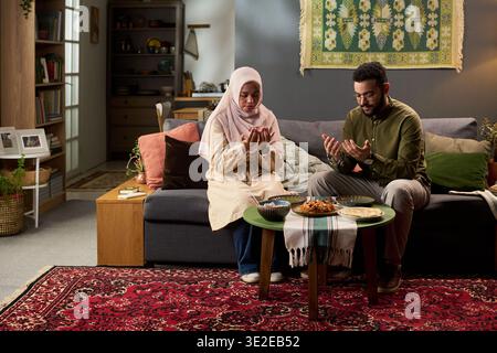 Young Muslim woman wearing hijab and young Muslim man sitting on sofa praying with hands raised in living room, traditional food on table in front of them Stock Photo