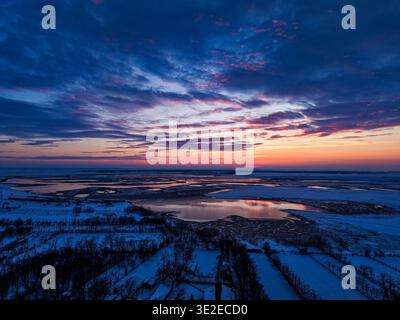 Drone photo of a sunset over a lake and a village covered by snow in Bucharest, Romania. Snow covered fields, frozen terrain, reflective water surface Stock Photo