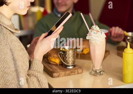 Caucasian young woman taking photo of milkshake with smartphone while sitting at table with Caucasian young man, both eating cheeseburgers in casual restaurant setting Stock Photo