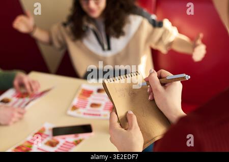 Unknown waitress writing order on notepad while speaking with two teenagers sitting at table, one gesturing with arms raised, restaurant menu and smartphone visible on table Stock Photo