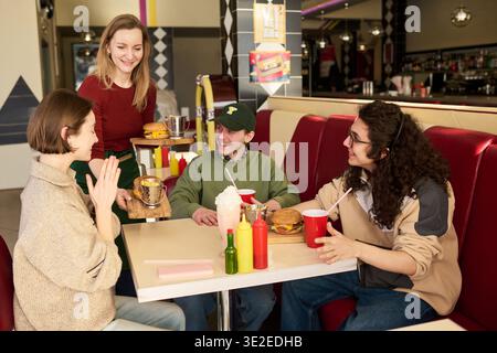 Group of young students sitting in diner booth smiling and talking while waitress serving burgers and drinks, ketchup and mustard bottles on table, milkshake and fries visible Stock Photo