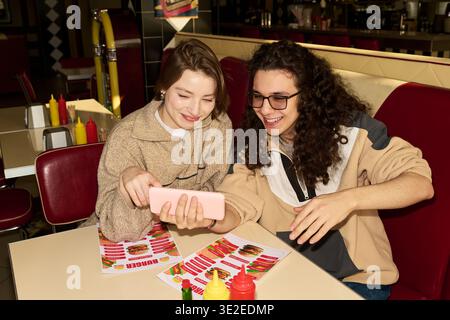 Caucasian young woman and Caucasian young man sitting at table in diner smiling and looking at smartphone together, holding menus with colorful food images, ketchup and mustard bottles visible Stock Photo