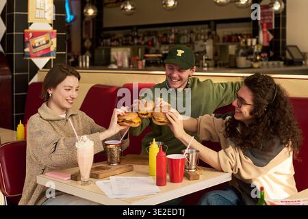 Three students sitting together in retro diner booth, smiling and holding burgers in hands, celebrating with milkshake and fries on table Stock Photo