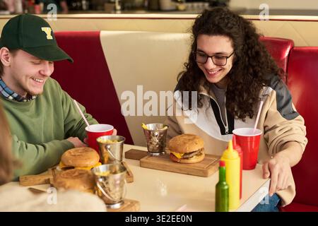 Two Caucasian young men sitting at table eating burgers and fries, smiling and talking, enjoying casual meal together in diner setting with drinks and condiments visible on table Stock Photo