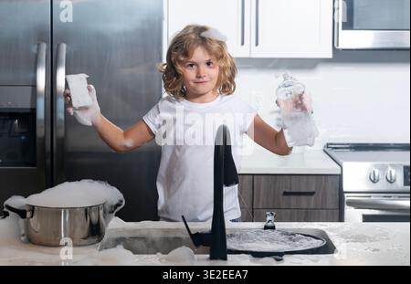 Boy washing dishes with bubbles. Child playing with foam in kitchen. Cute kid cleaning dishes at home. Happy child splashing soap foam. Funny boy Stock Photo