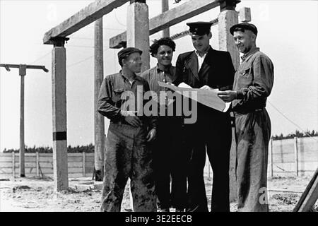1969, Slavyansk railway depot. Engaging vintage black and white archival photograph depicting a collaborative moment at a railway construction site. A supervisor wearing an official uniform and cap is actively discussing a large paper blueprint with three attentive workers dressed in dirty work clothes. They stand near large concrete pillars intended for the electrified track system. This scene perfectly illustrates industrial teamwork and meticulous planning, representing life before the war, peaceful Donbas. Stock Photo