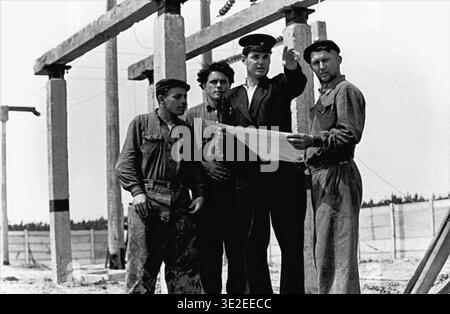 1969, Slavyansk railway depot. Engaging vintage black and white archival photograph depicting a collaborative moment at a railway construction site. A supervisor wearing an official uniform and cap is actively discussing a large paper blueprint with three attentive workers dressed in dirty work clothes. They stand near large concrete pillars intended for the electrified track system. This scene perfectly illustrates industrial teamwork and meticulous planning, representing life before the war, peaceful Donbas. Stock Photo