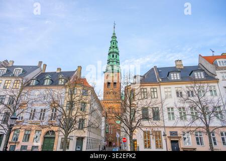People walk around the historic Nikolaj Kunsthal, a contemporary arts center in the former St. Nicholas Church. The tall tower stands among nearby buildings in the city. Stock Photo