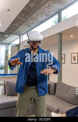 Bearded man wearing VR headset and headphones, reaching forward in living room near pool table Stock Photo