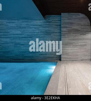 Indoor swimming pool with blue underwater lighting. Minimalist spa interior with illuminated blue pool water, textured stone walls, and wooden ceiling. Stock Photo