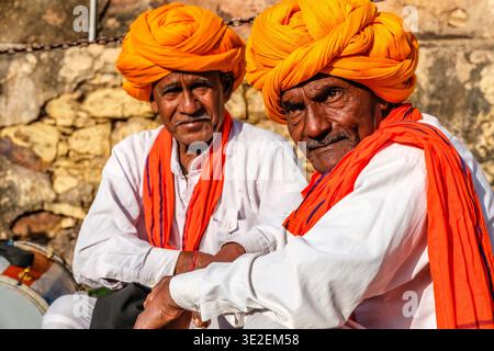 Two men wearing orange turbans and traditional attire sit together during the Bundi Utsav festival in front of a weathered stone wall. Stock Photo