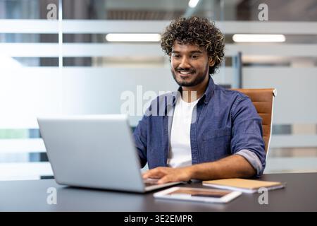 Indian smiling young man sitting in the office at the desk and working, studying on a laptop. Stock Photo