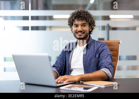Portrait of a young Indian man sitting at a desk in the office, working on a laptop and smiling, looking at the camera. Stock Photo