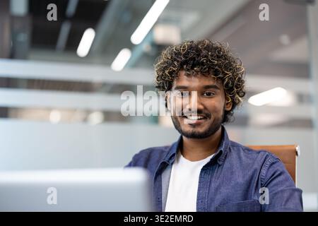Close-up photo of a smiling young Indian man in an office working on a laptop. Stock Photo