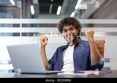 Portrait of a happy young Indian man sitting in the office at a table with a laptop, raising his hands up and rejoicing. Stock Photo