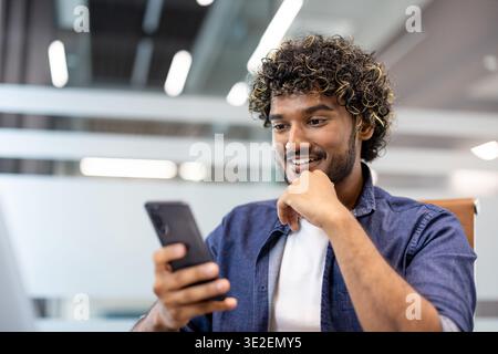 Close-up photo of a young Indian man working in an office sitting at a desk and smiling while using his mobile phone. Stock Photo