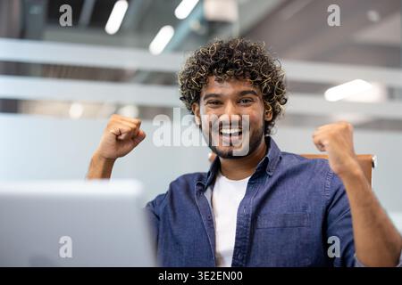 Close-up portrait of a young Indian man sitting in an office with a laptop and looking at the camera. He raised his hands up, showing a sign of strength. Stock Photo