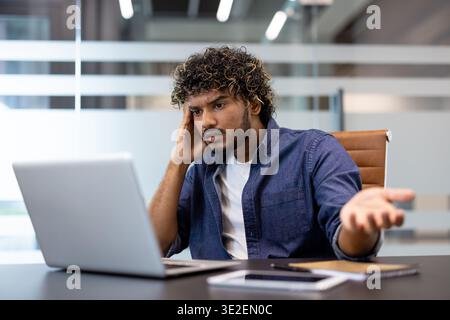 Close-up photo of a young Indian man sitting at an office desk, looking angrily at the laptop screen and having an emotional conversation online. Stock Photo