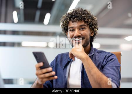 Close-up portrait of a young Indian man sitting in a chair at a desk in an office, holding a phone and smiling at the camera. Stock Photo