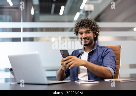 Portrait of a smiling young Indian man sitting at an office desk, holding a phone in his hand and smiling at the camera. Stock Photo