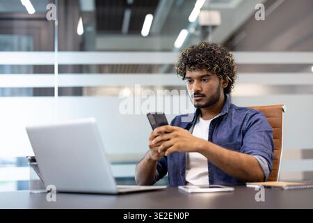 Serious young Indian man sitting in the lobby at his desk and using his mobile phone. Stock Photo