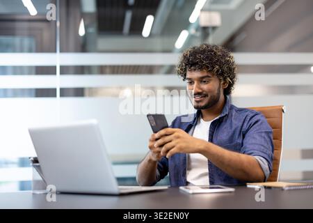 Smiling young Indian man working in an office, sitting at a desk and using a mobile phone. Stock Photo
