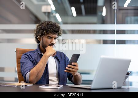 A serious and thoughtful young Indian man is sitting at his desk in the office and looking thoughtfully at his mobile phone screen. Stock Photo
