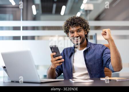 Happy young Indian man sitting at office desk with laptop, holding phone in hand and showing success and victory sign to camera. Stock Photo