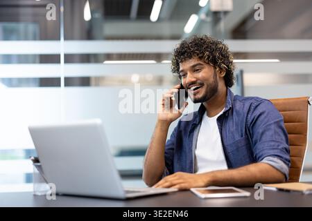 A young Indian man is sitting in an office at a desk in front of a laptop and talking on the phone with a smile. Stock Photo
