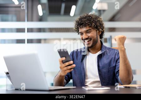 Happy young Indian man sitting in the office at his desk, looking at the mobile phone screen and rejoicing at the news and success, showing a victory gesture with his hand. Stock Photo