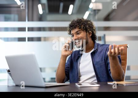 A young Indian man is sitting at his desk in the office and smilingly talking on the phone. Stock Photo